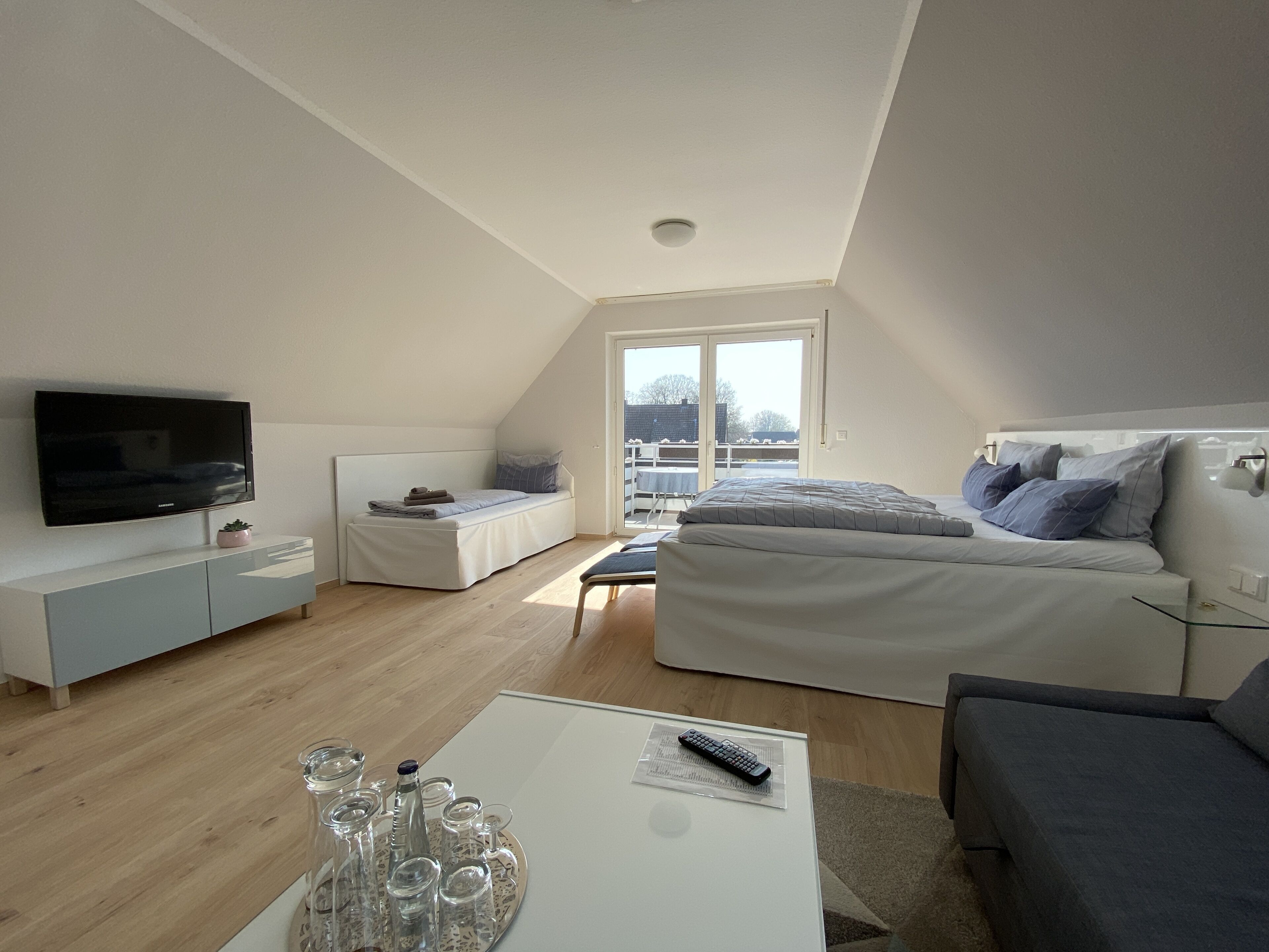 A modern, well-lit room with a flat-screen TV, a sofa, a bed, and a balcony with a view.
