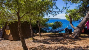 A hammock between trees by a clear blue sea, with a large rock structure and a tent in the background. - Cast Away - Eco Village (Jelsa)