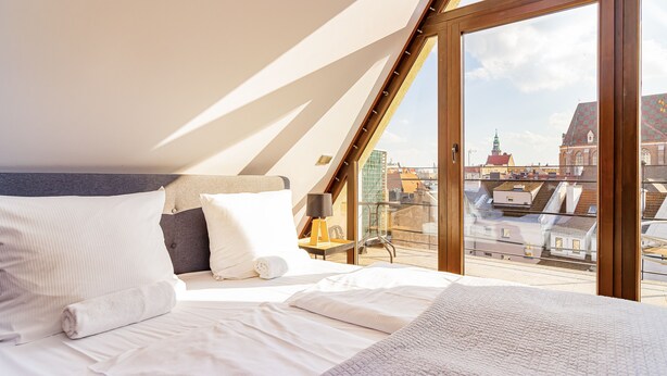 A bedroom with a large bed, a nightstand, a lamp, and a view of the city through a glass door.