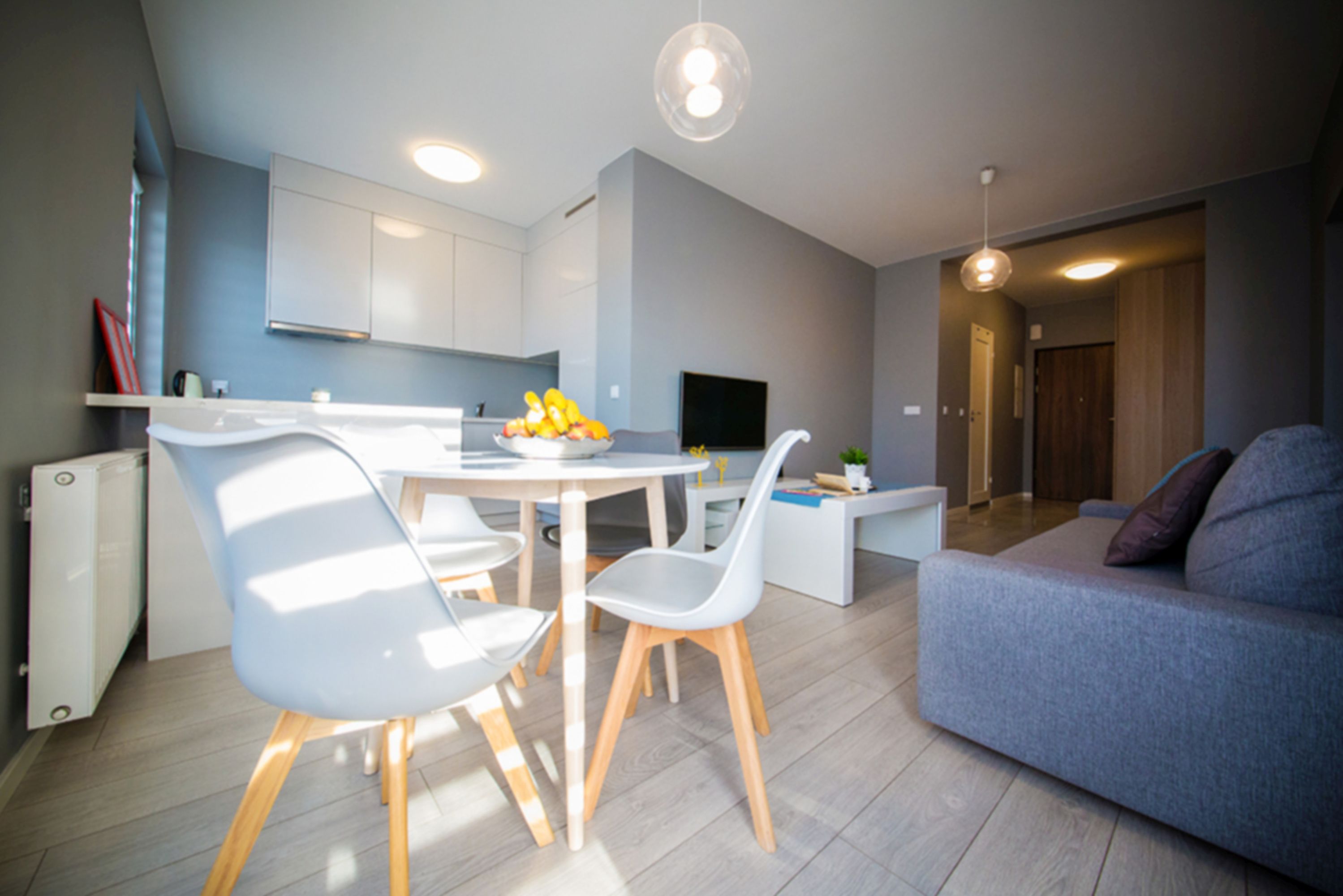 Una sala de estar moderna con un sofá gris, una mesa de comedor con sillas y una zona de cocina con armarios y un televisor.