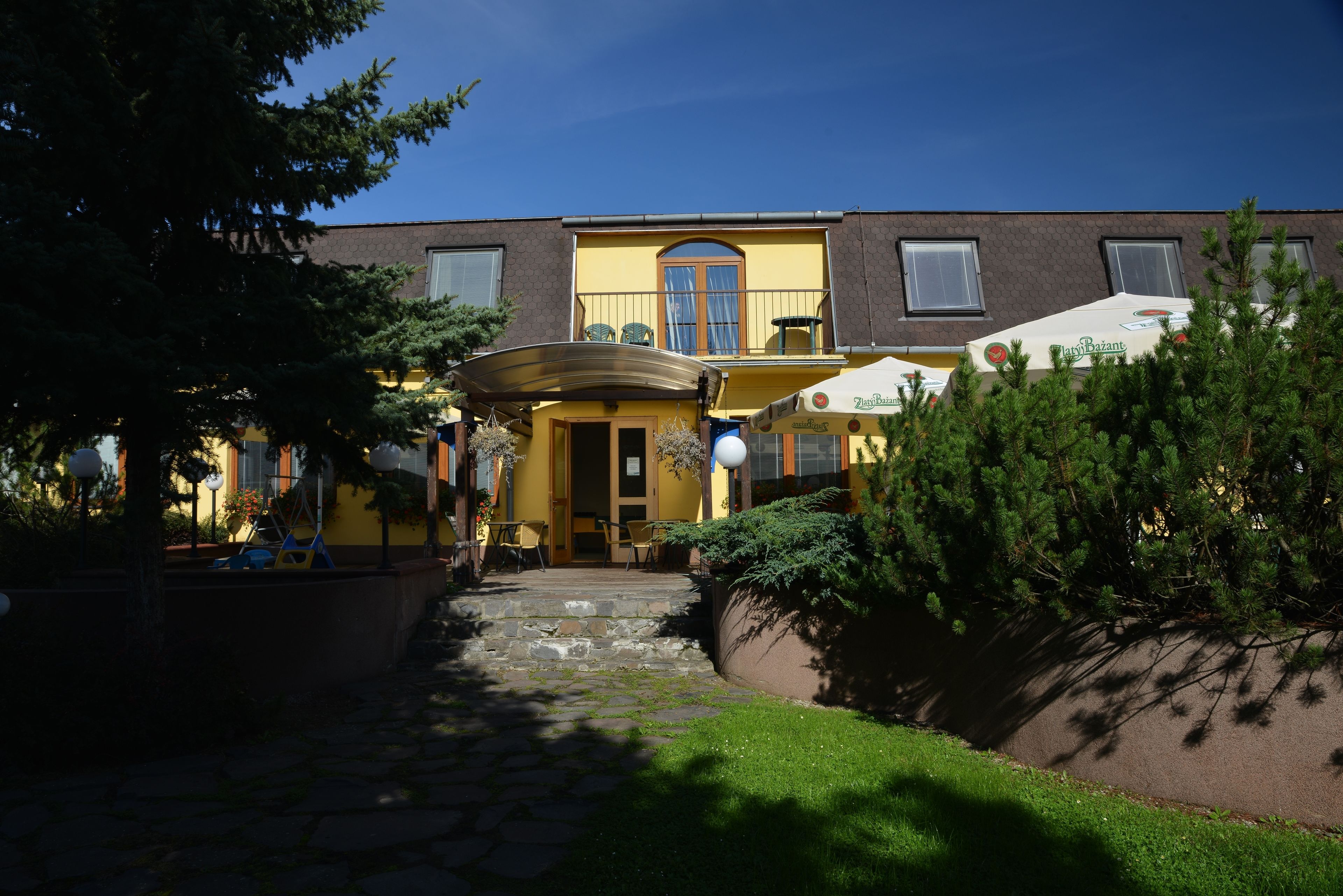 A building with a yellow facade, a stone pathway leading to the entrance, outdoor seating with umbrellas, and a small garden area.
