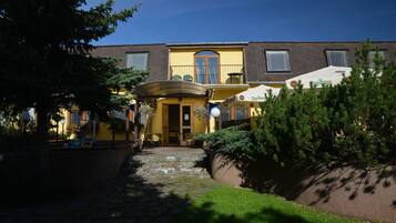 A building with a yellow facade, a stone pathway leading to the entrance, outdoor seating with umbrellas, and a small garden area.