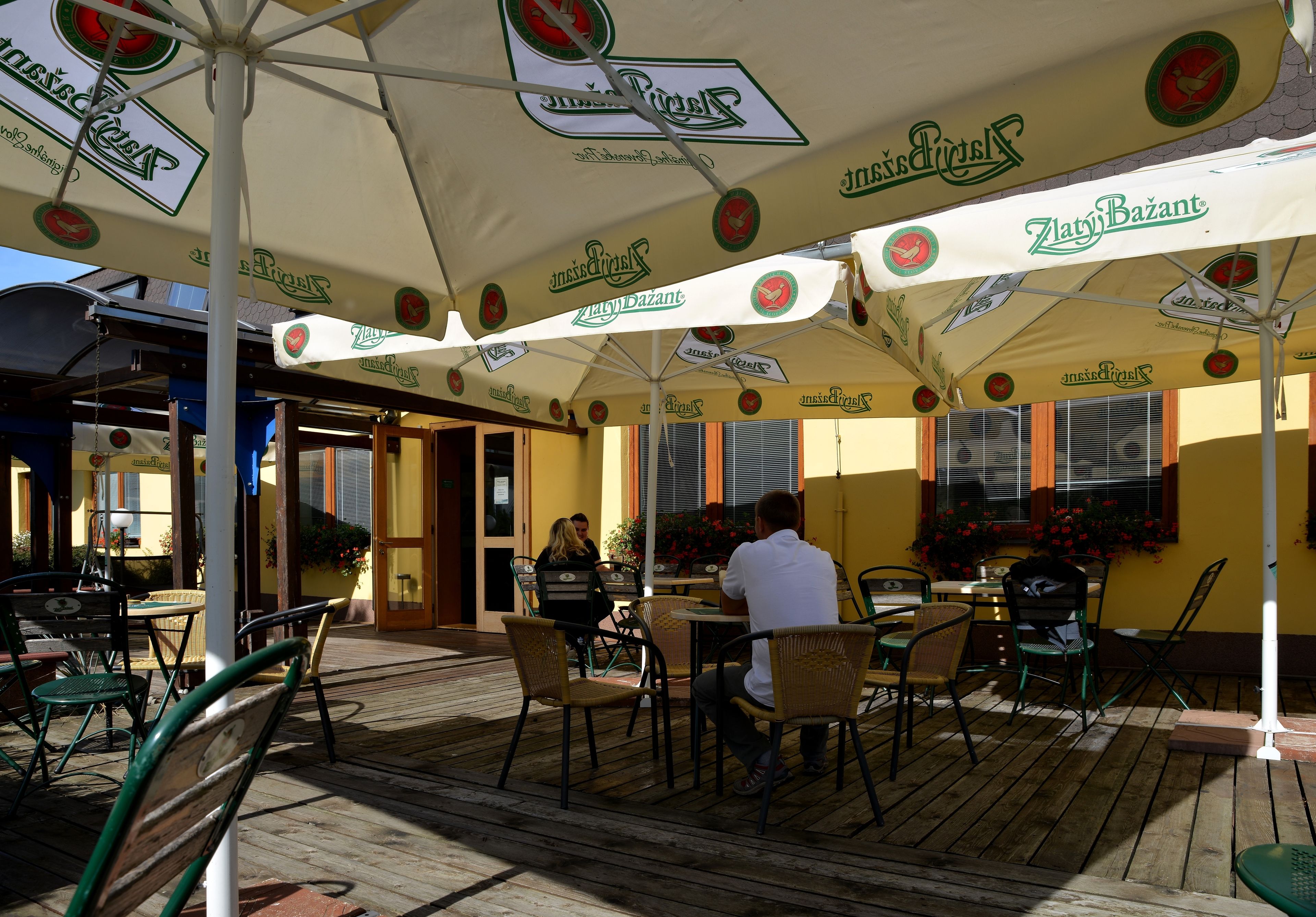 Un café al aire libre con mesas y sillas bajo sombrillas con logotipos, una terraza de madera y una pared amarilla con ventanas.