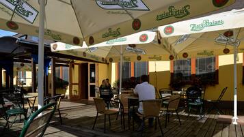 Un café al aire libre con mesas y sillas bajo sombrillas con logotipos, una terraza de madera y una pared amarilla con ventanas.