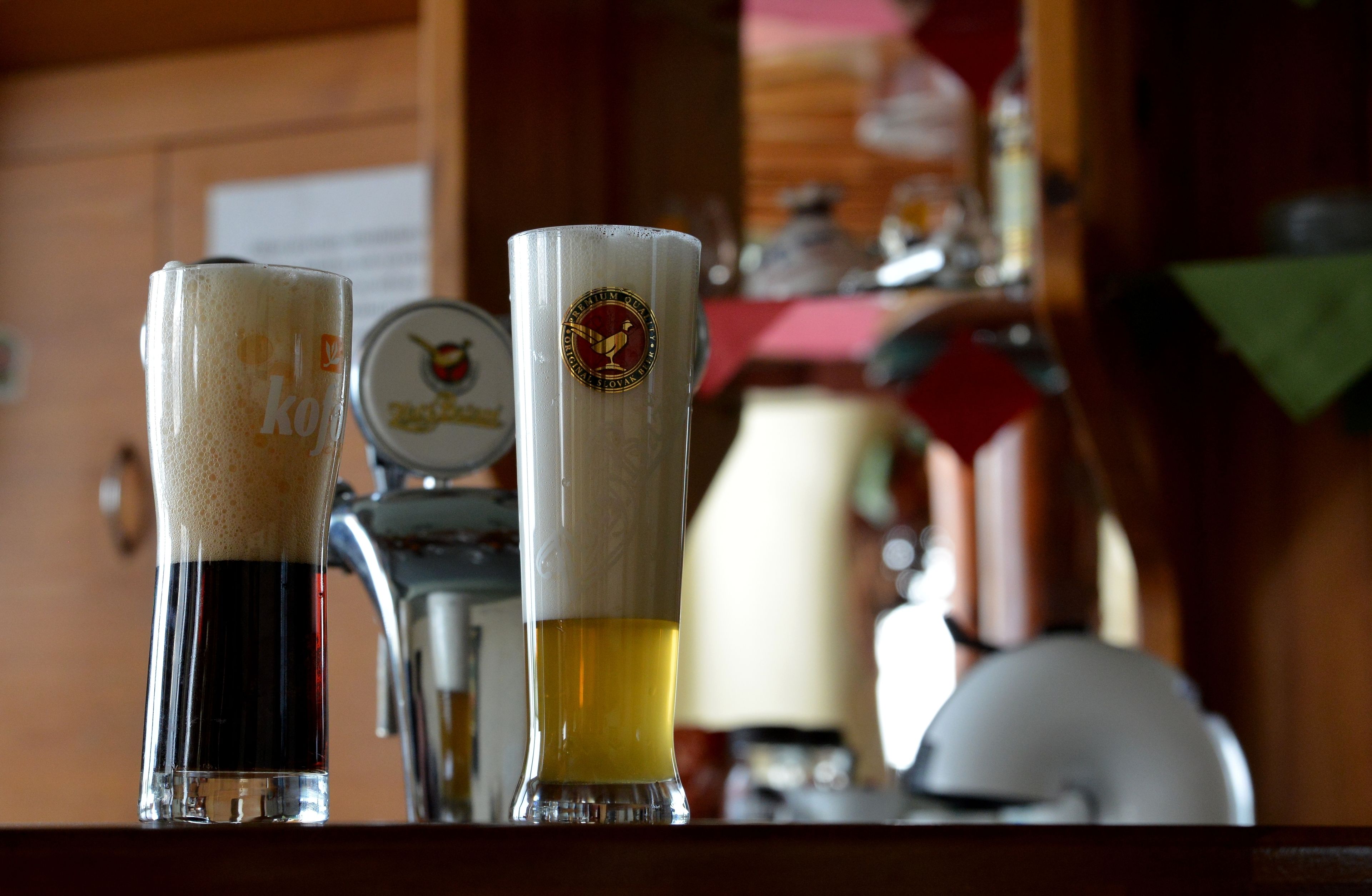 Two beer glasses on a bar counter with a tap and a beer keg in the background.