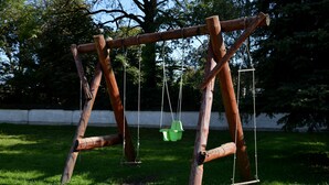 Un conjunto de columpios de madera en un parque con césped y árboles al fondo.