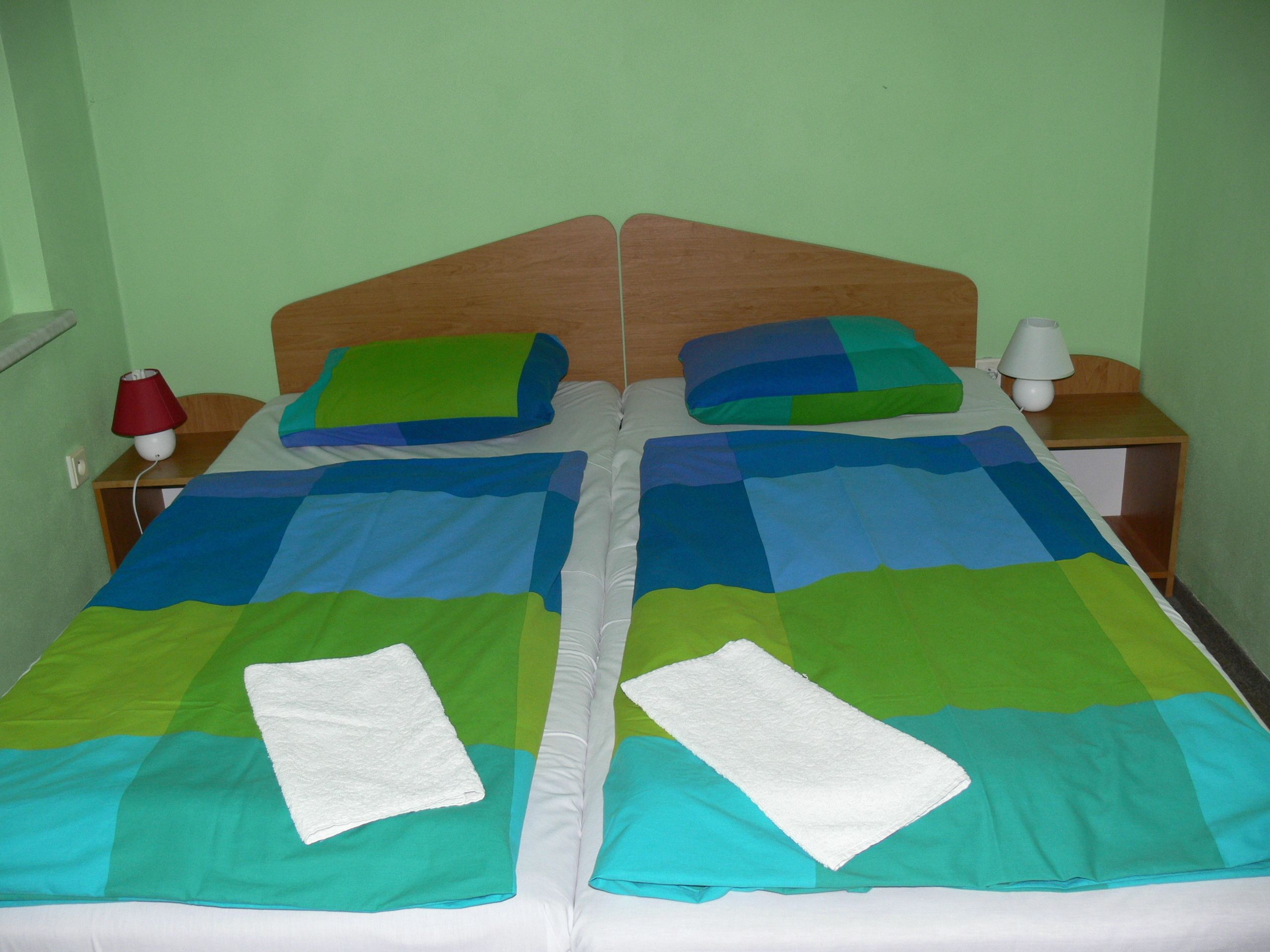 A double bed with a green and blue quilt, two pillows, a white towel, and a bedside table with a red lamp.