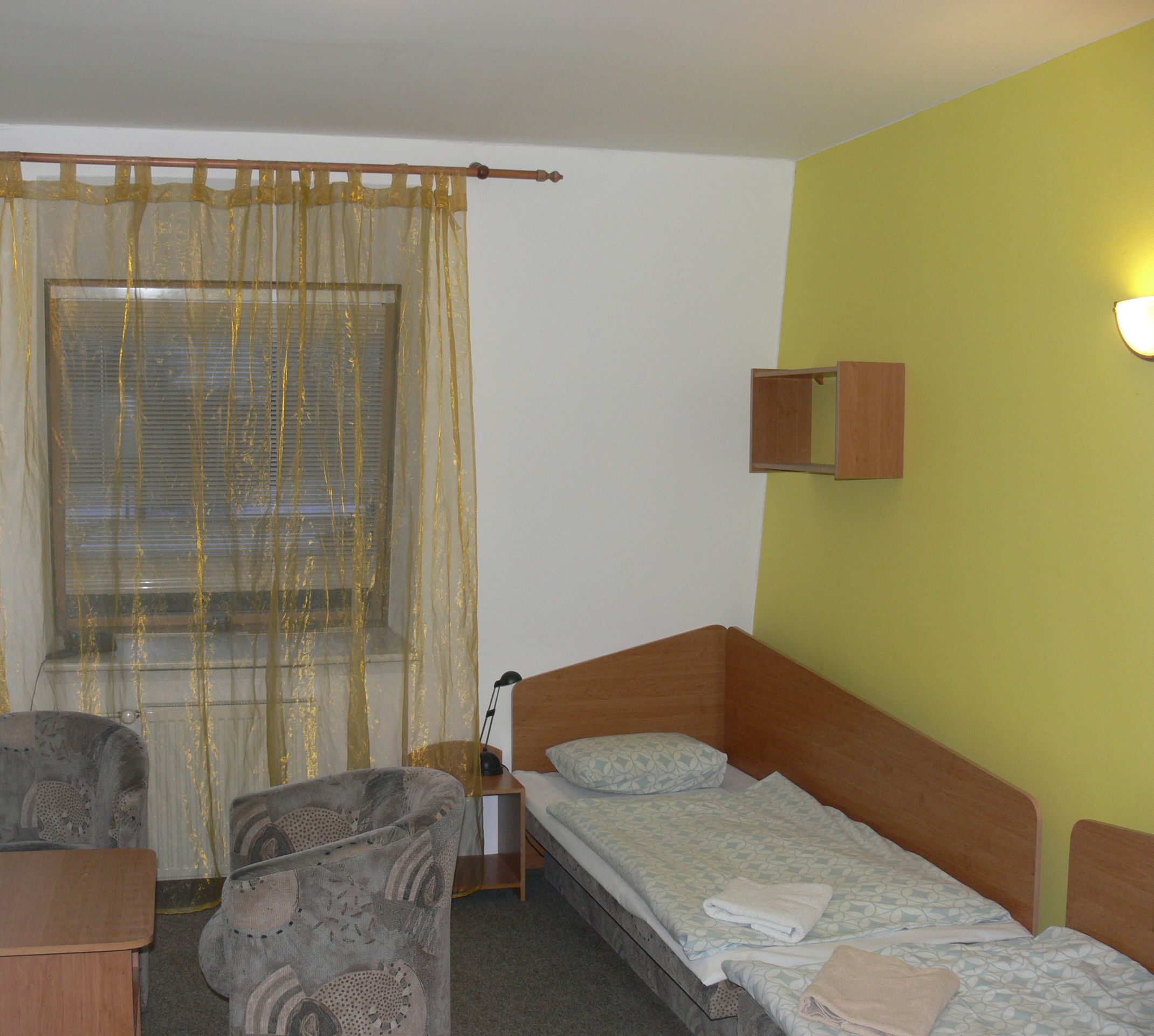A small, single-room accommodation with a bed, a chair, a small table, and a window with curtains.
