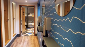 Un couloir d’hôtel avec un sol à carreaux, un miroir à motifs en zigzag et un panneau indiquant la présence d’une douche sur une porte.