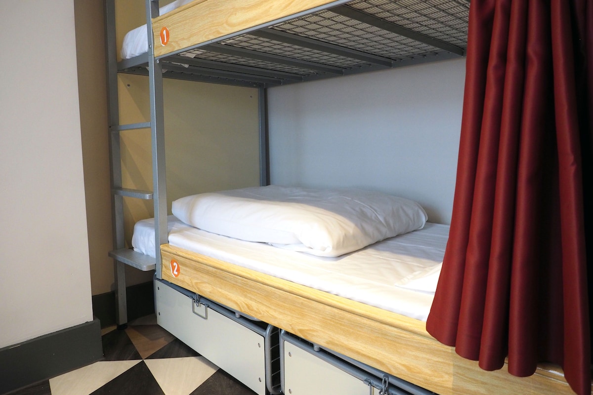 Una cama litera con estructura de madera, escalera de metal y cortina roja.