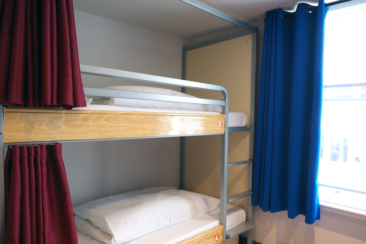 Ein Schlafraum mit Etagenbetten, einem Fenster, Vorhängen und einer Holzkiste.