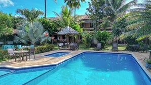 A swimming pool surrounded by lounge chairs and tables, with a thatched-roof structure and lush greenery in the background. - Pousada Saudosa Maloca (Porto Seguro)