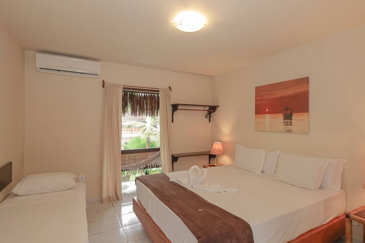 A hotel room with a bed, a nightstand, a lamp, a painting, and an air conditioning unit.