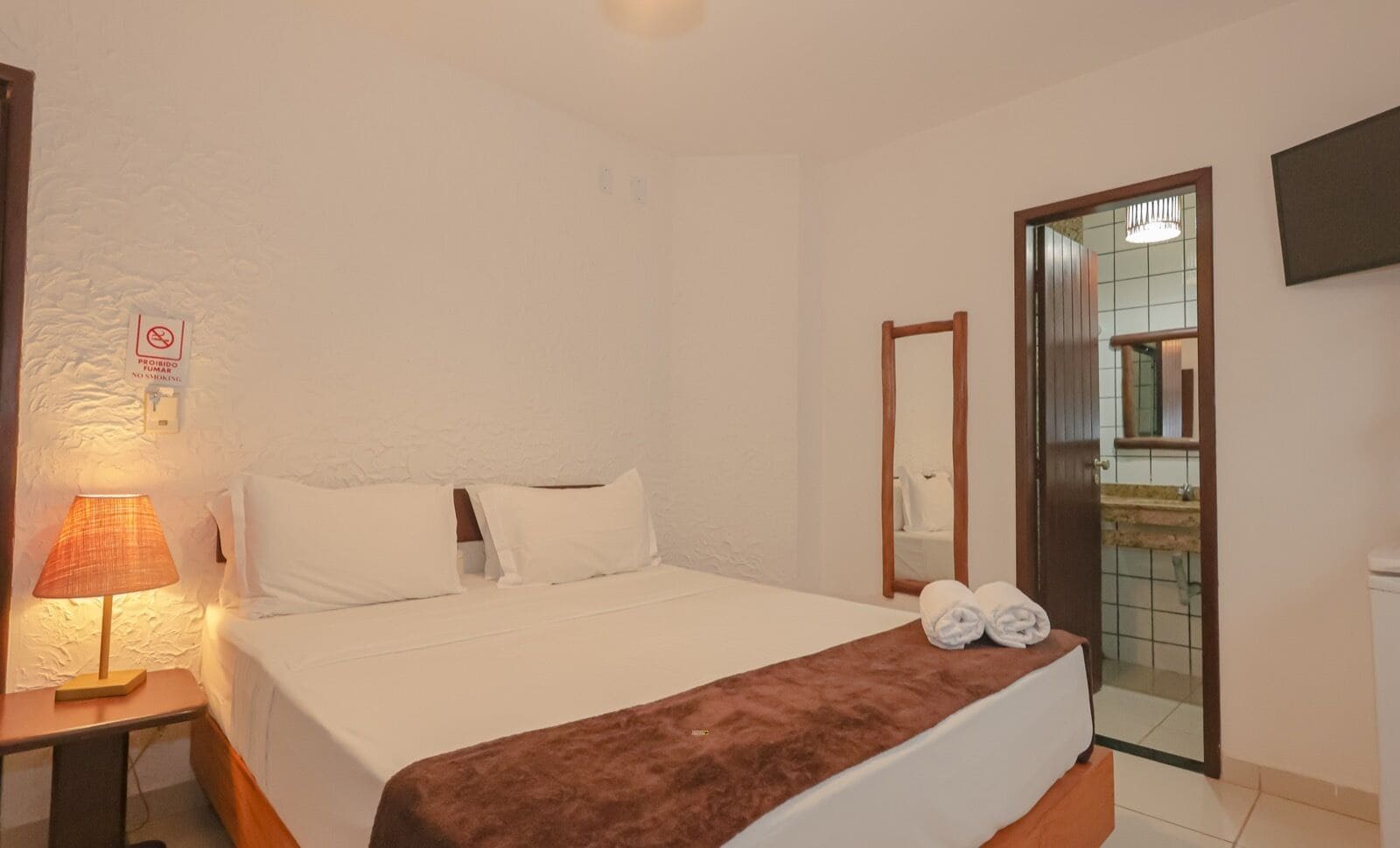 A hotel room with a bed, a nightstand with a lamp, a bathroom visible through an open door, and a sign prohibiting smoking.