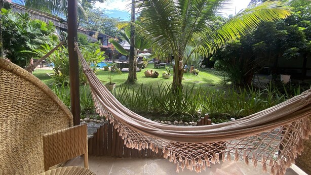 A hammock in a lush garden with a pool and outdoor furniture. - Pousada Saudosa Maloca (Porto Seguro)