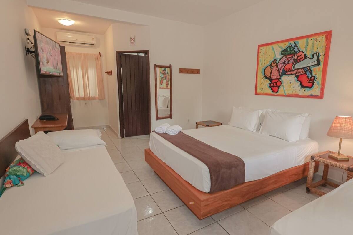 A hotel room with a large bed, two smaller beds, a bedside table with a lamp, a painting on the wall, and a door leading to another room.
