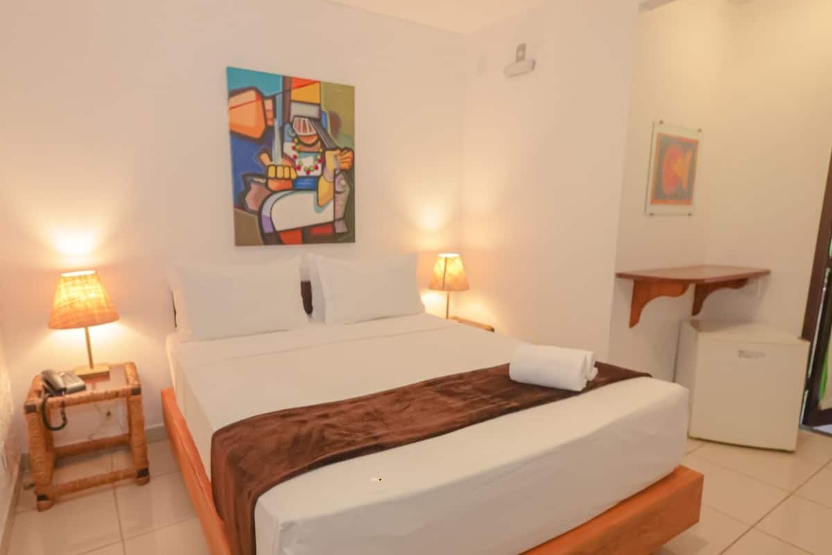 A neatly arranged hotel room with a large bed, two bedside lamps, a small bedside table with a lamp, a wall-mounted painting, and a small cabinet.