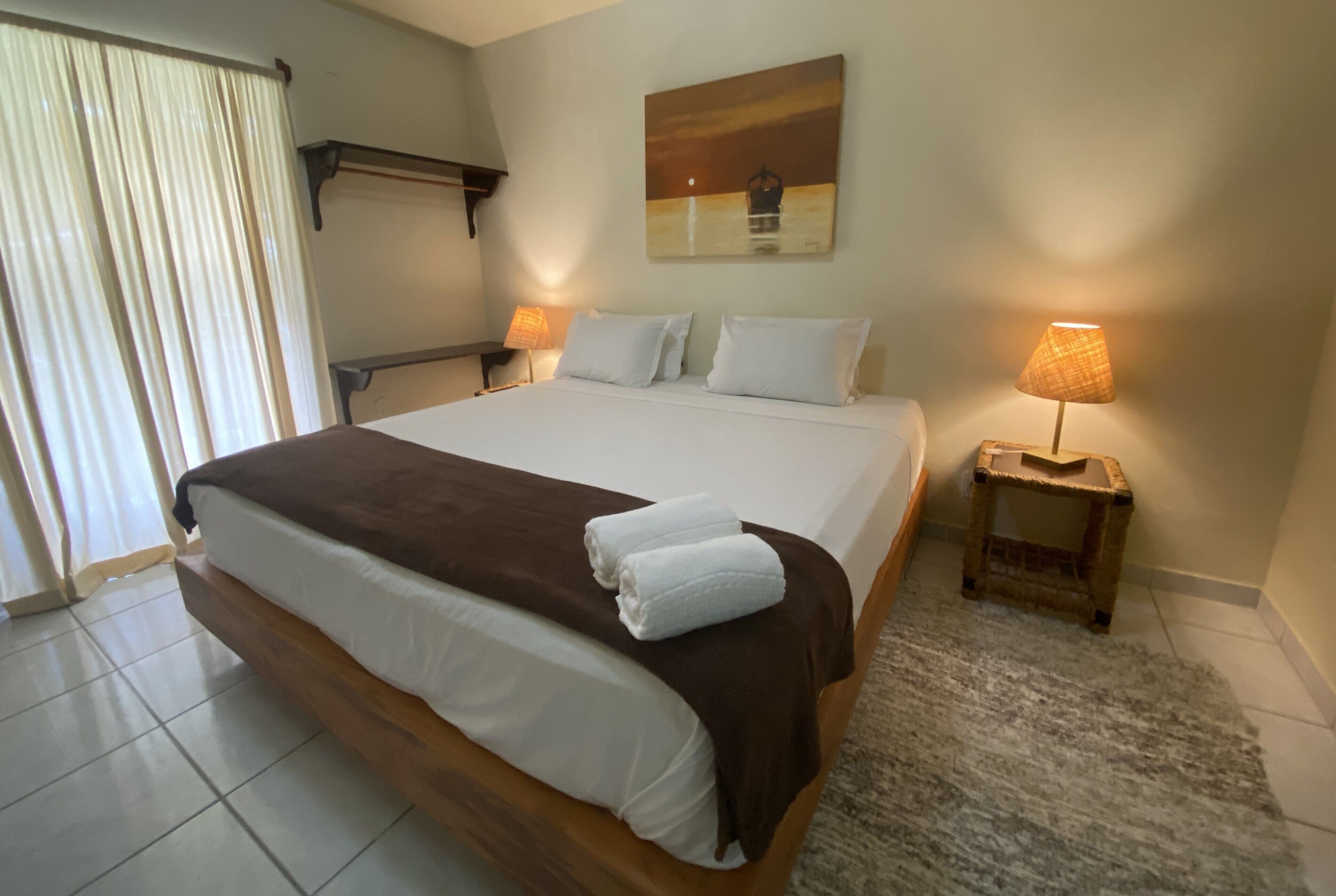 A neatly made bed with white and brown linens, two bedside lamps, a painting on the wall, and a small rug on the floor.
