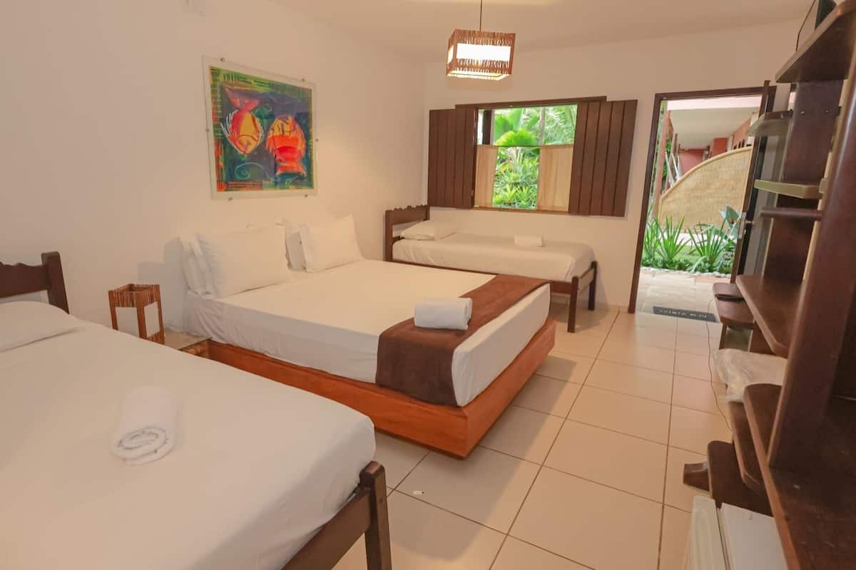 A hotel room with three beds, a large window with curtains, a painting on the wall, and a view of greenery outside.