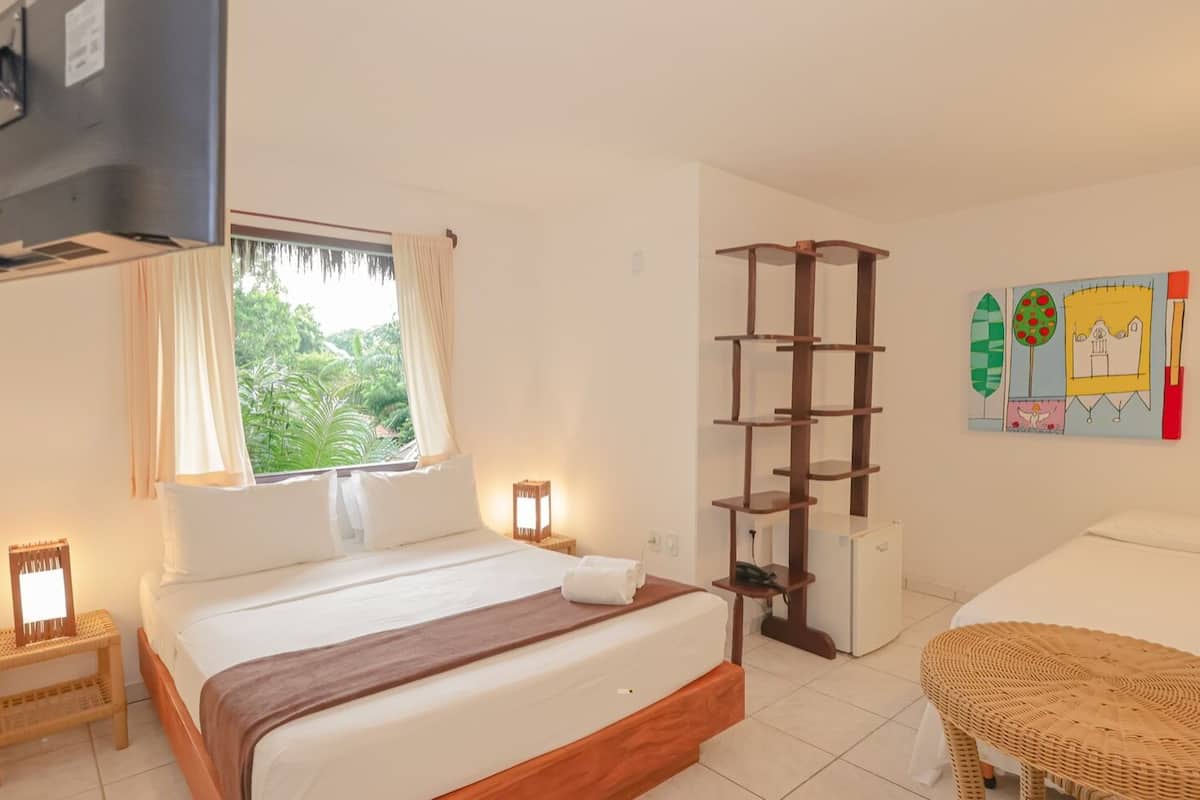 A hotel room with a bed, bedside tables, a small refrigerator, a wicker chair, and a wall with a colorful painting.