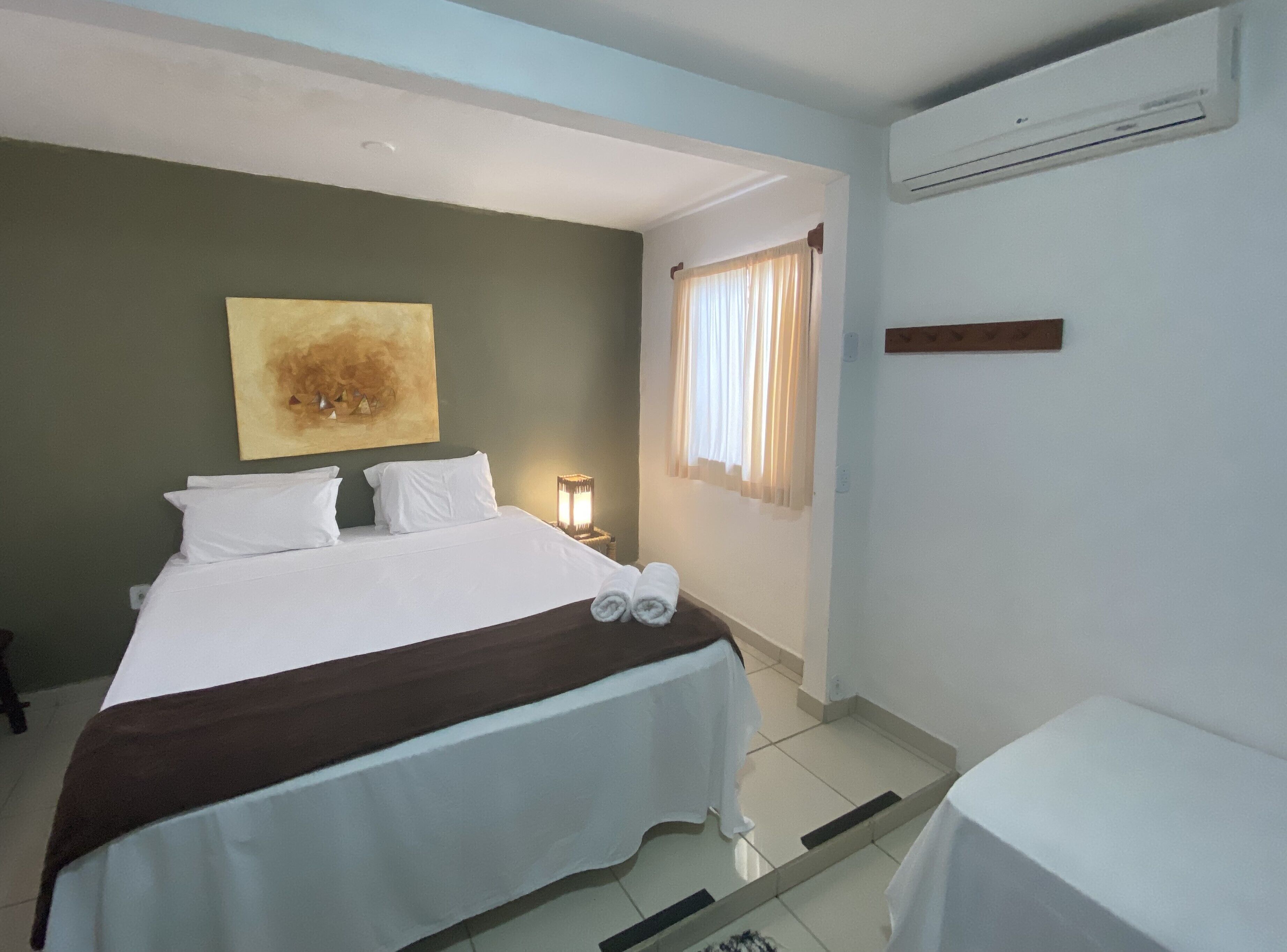 A hotel room with a bed, a nightstand, a lamp, a window with curtains, and an air conditioning unit.