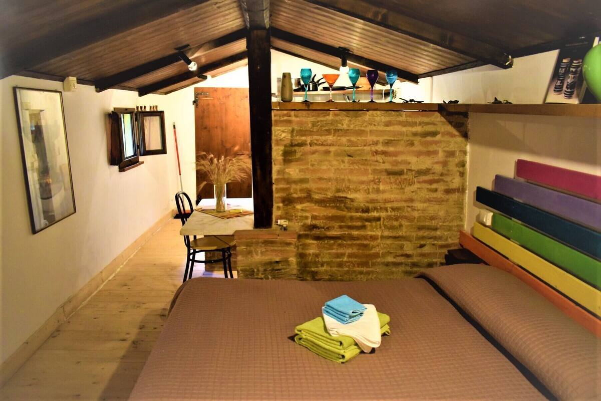 A cozy room with a wooden ceiling, a bed with a brown mattress, a small table with a vase of dried plants, and a colorful bench.