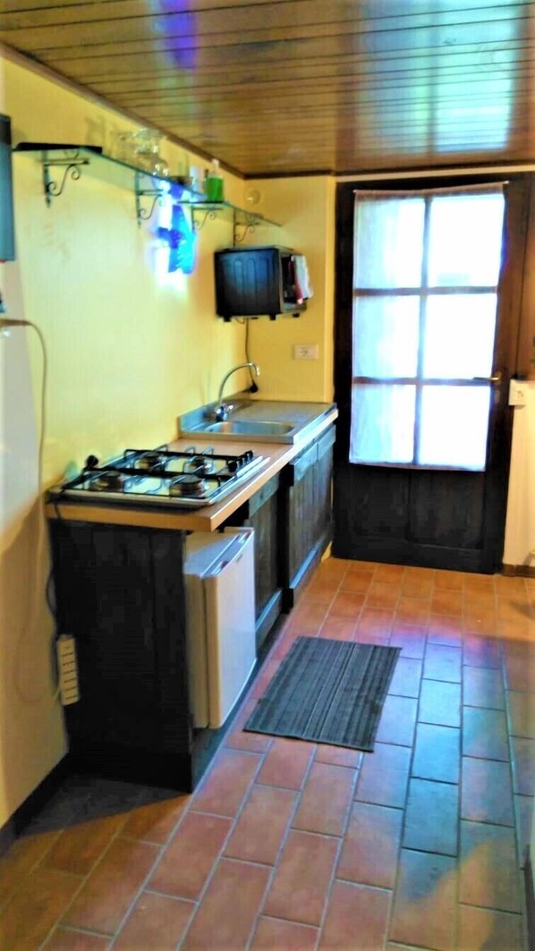 A compact kitchen with a gas stove, a sink, and a small refrigerator. There is a window with blinds, and the floor is tiled. - Casale Civetta (Trecastelli)