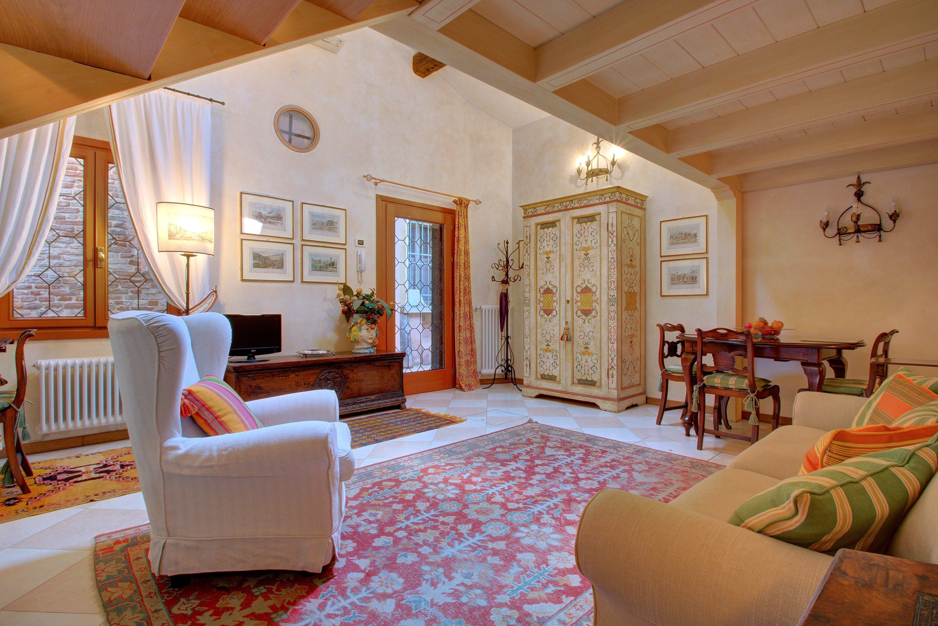 Una sala de estar acogedora con un sillón blanco, un sofá, una mesa de comedor con sillas, un aparador y un amplio ventanal con cortinas.