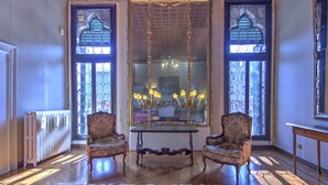 Une pièce dotée de deux fauteuils richement décorés, d’une table surmontée d’un lustre et de grandes fenêtres aux motifs complexes.