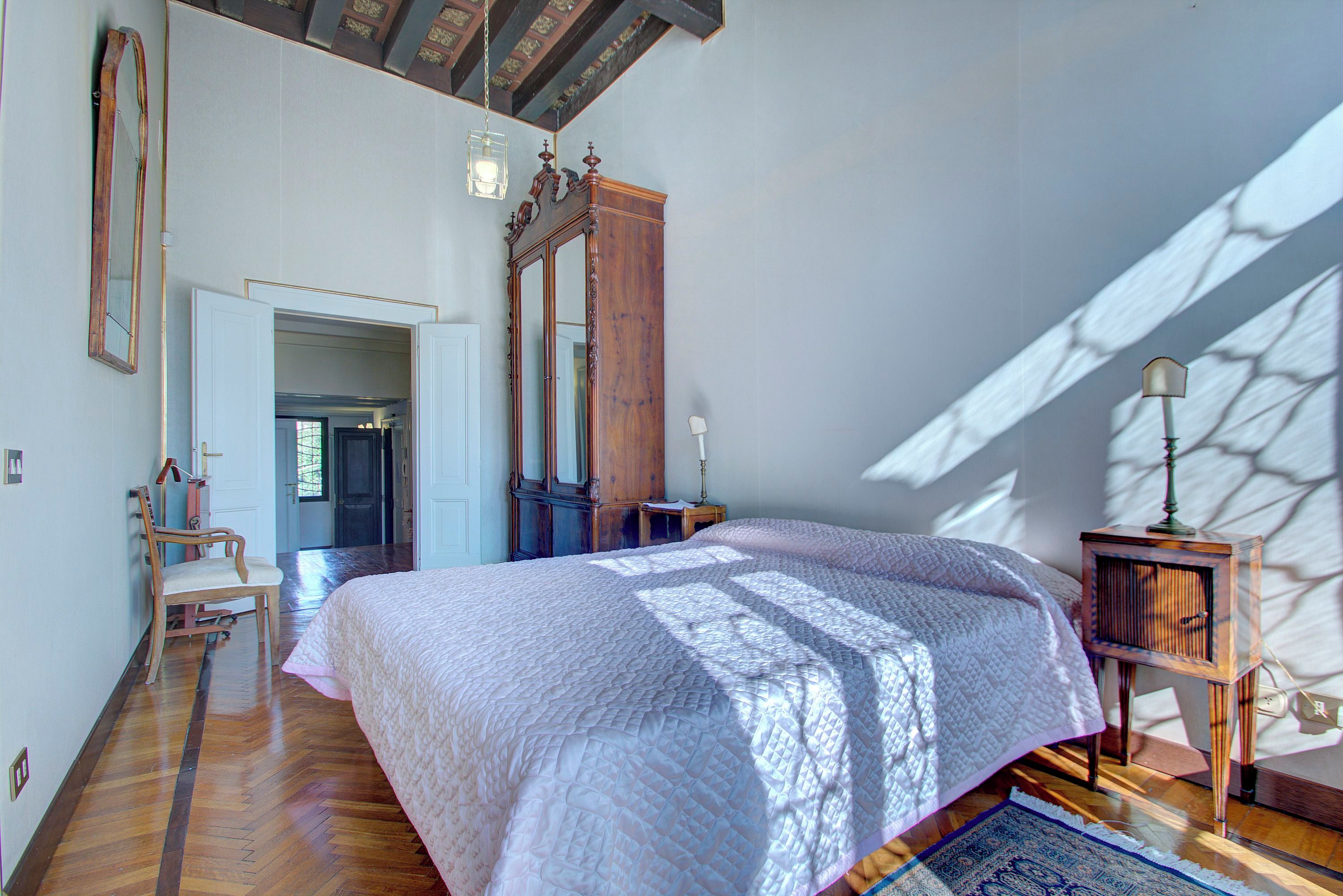 Une chambre à coucher avec un grand lit, un plancher en bois, un miroir, une chaise et une table de chevet avec une lampe.