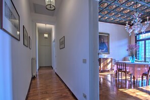 Un pasillo con suelo de madera, paredes blancas y cuadros enmarcados. Hay un comedor con mesa y sillas, un candelabro y una ventana con vistas.