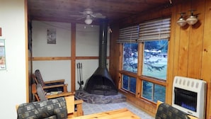 A cozy cabin interior with a stone fireplace, wooden walls, and a large window offering a view of snow-covered trees. - Woman River Camp (Unorganized Kenora District)