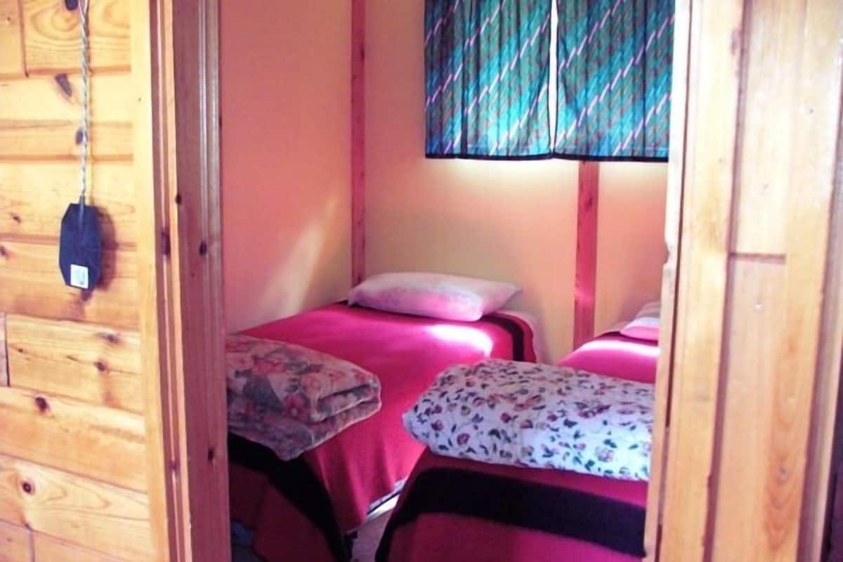 Um quarto pequeno e acolhedor com duas camas, cada uma com uma colcha florida e um travesseiro. Uma porta de madeira é visível à esquerda, e uma janela com cortina colorida à direita.