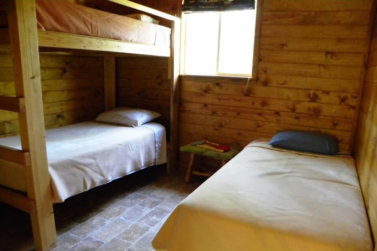 Uma cabana com duas beliches e uma cama de solteiro, paredes de madeira e uma janela.