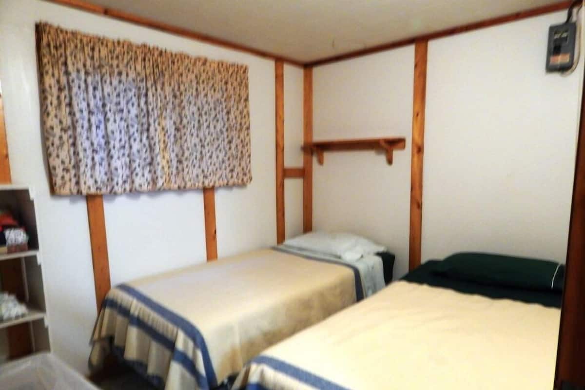 Quarto pequeno e simples com duas camas de solteiro, uma prateleira de madeira e uma cortina.