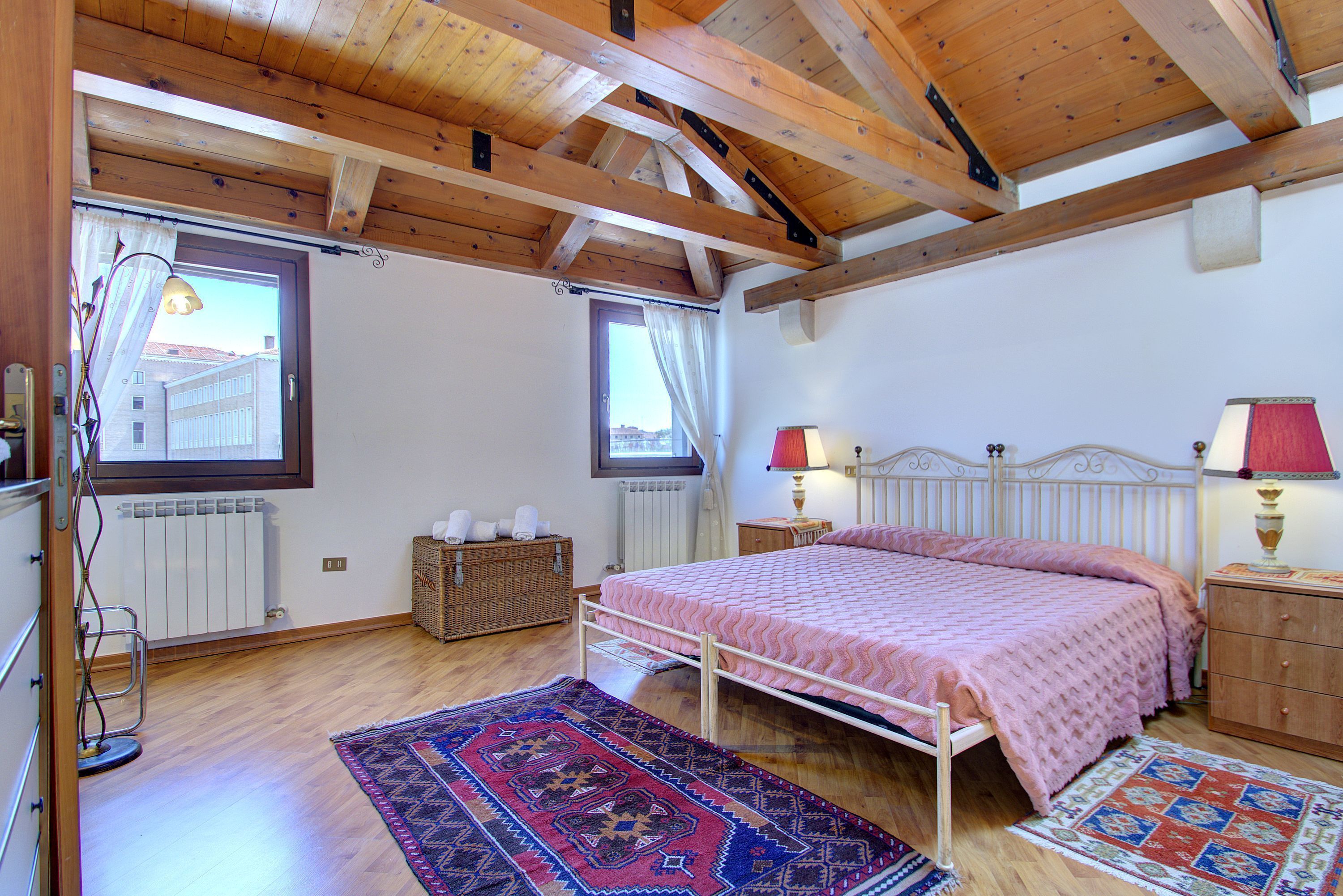 A bedroom with a wooden ceiling, a bed with a pink bedspread, two bedside lamps, a wooden dresser, and a patterned rug.