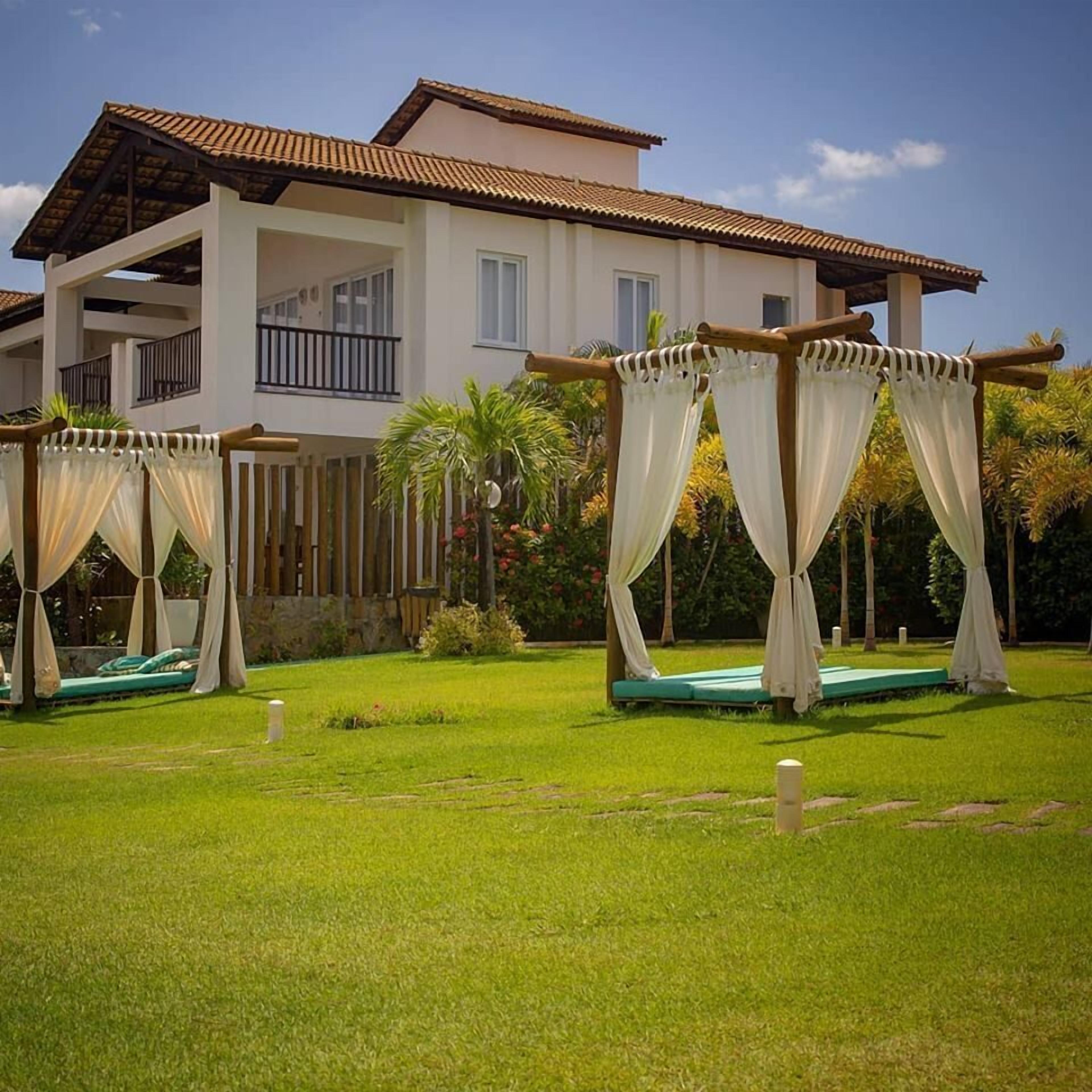 A two-story house with a balcony, a garden with a lawn, and a pergola with white curtains.