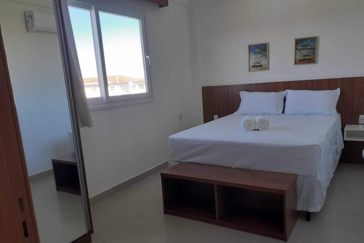 A bedroom with a bed, a bedside table, a nightstand, a bench, a window, and two wall-mounted pictures.