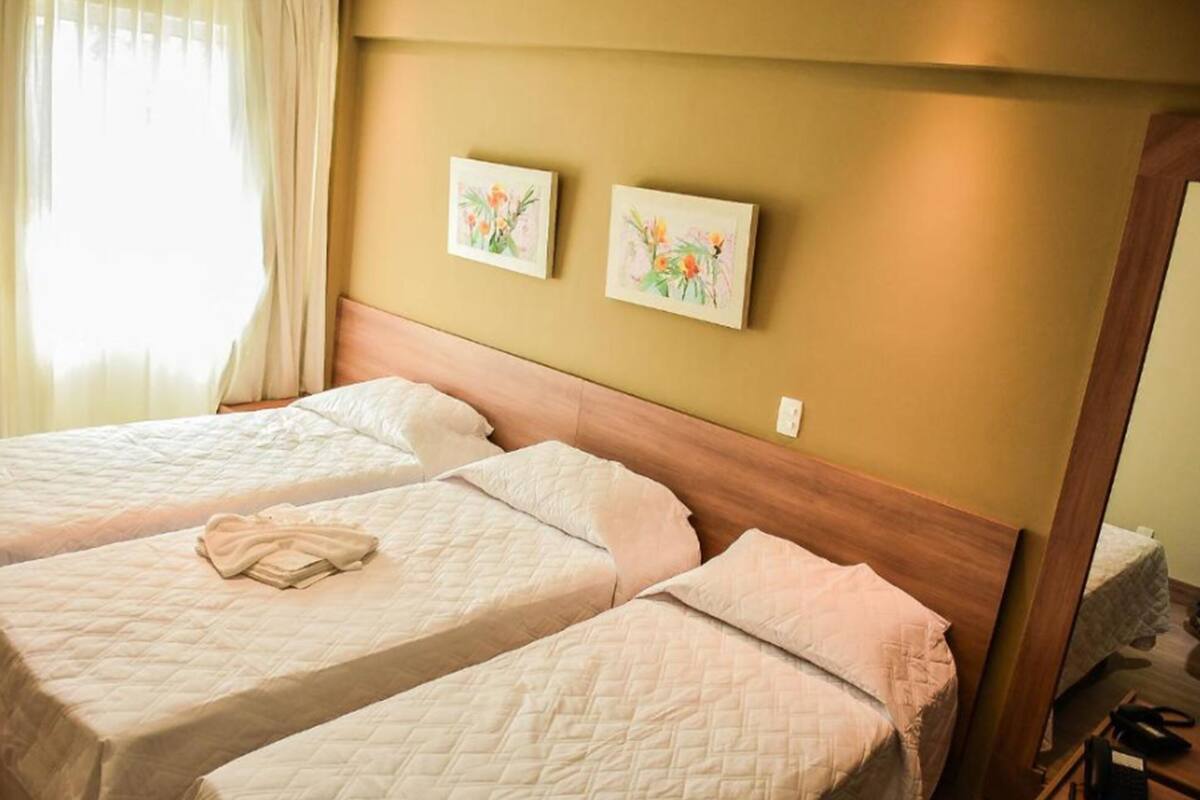 A hotel room with two beds, beige bedding, a wooden headboard, and two floral paintings on the wall.