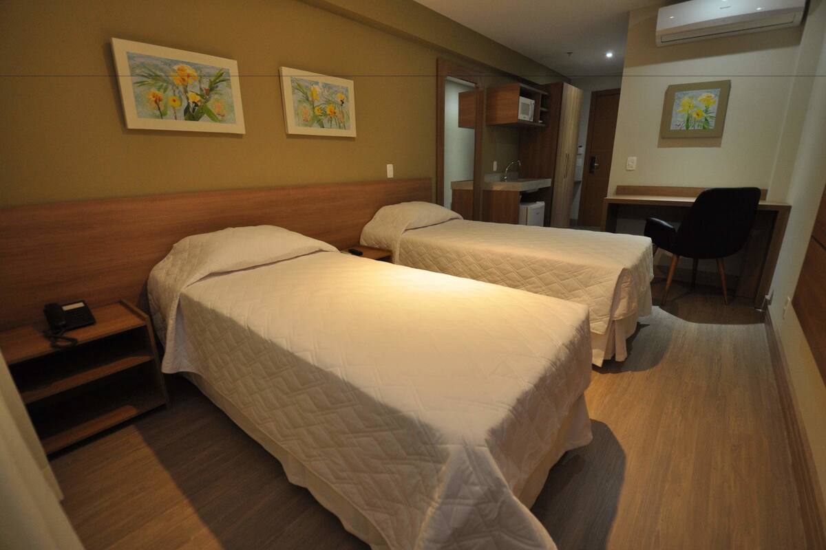 A hotel room with two beds, a desk, a chair, and a small kitchenette.