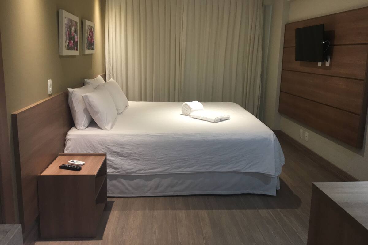 A neatly made bed with white linens, a wooden headboard, a bedside table with a remote control, and a flat-screen TV mounted on the wall.