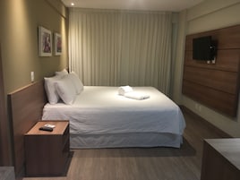 A neatly made bed with white linens, a wooden headboard, a bedside table with a remote control, and a flat-screen TV mounted on the wall.