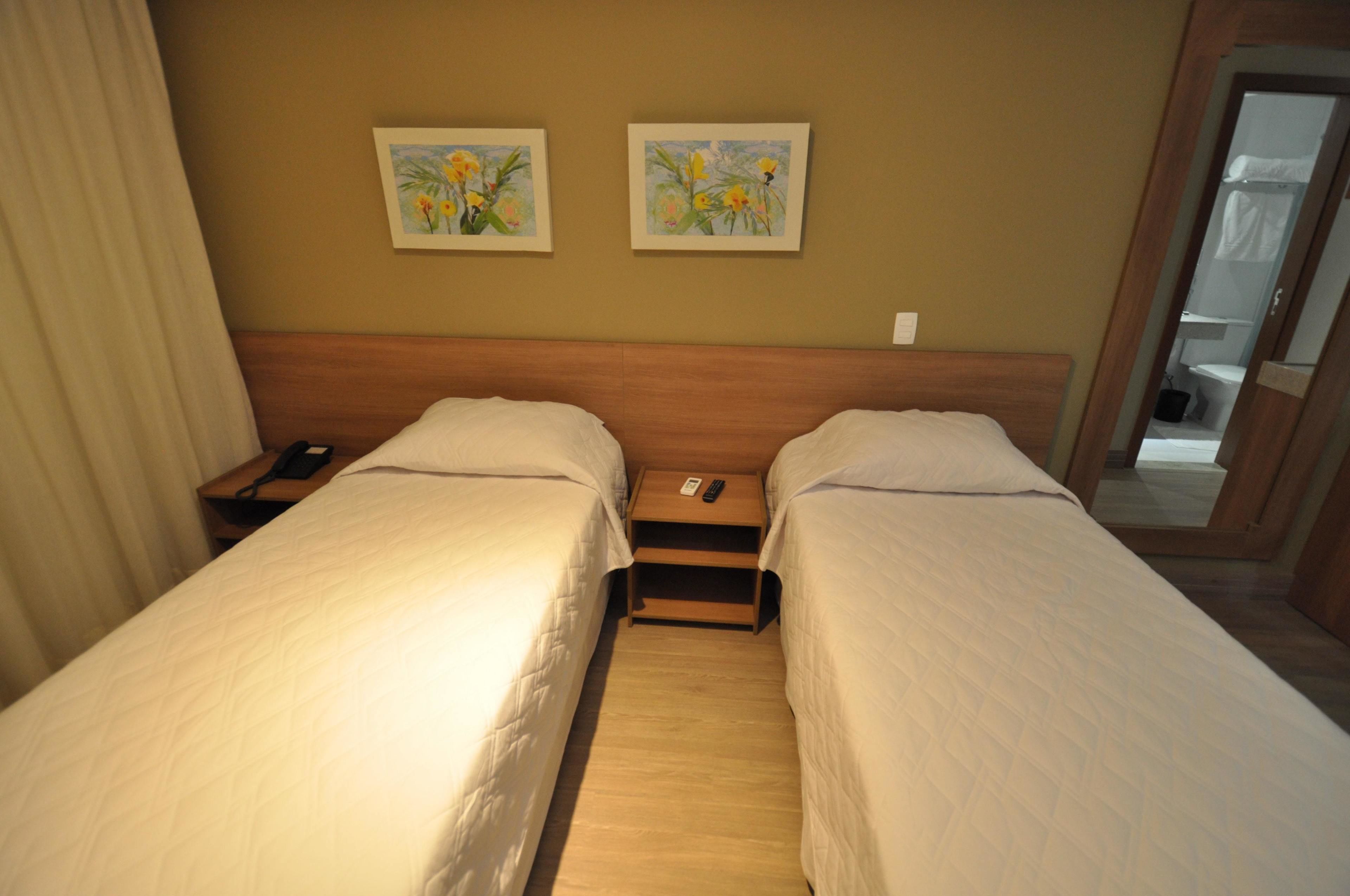 A hotel room with two single beds, a wooden headboard, a nightstand with a phone, and two framed floral paintings on the wall.