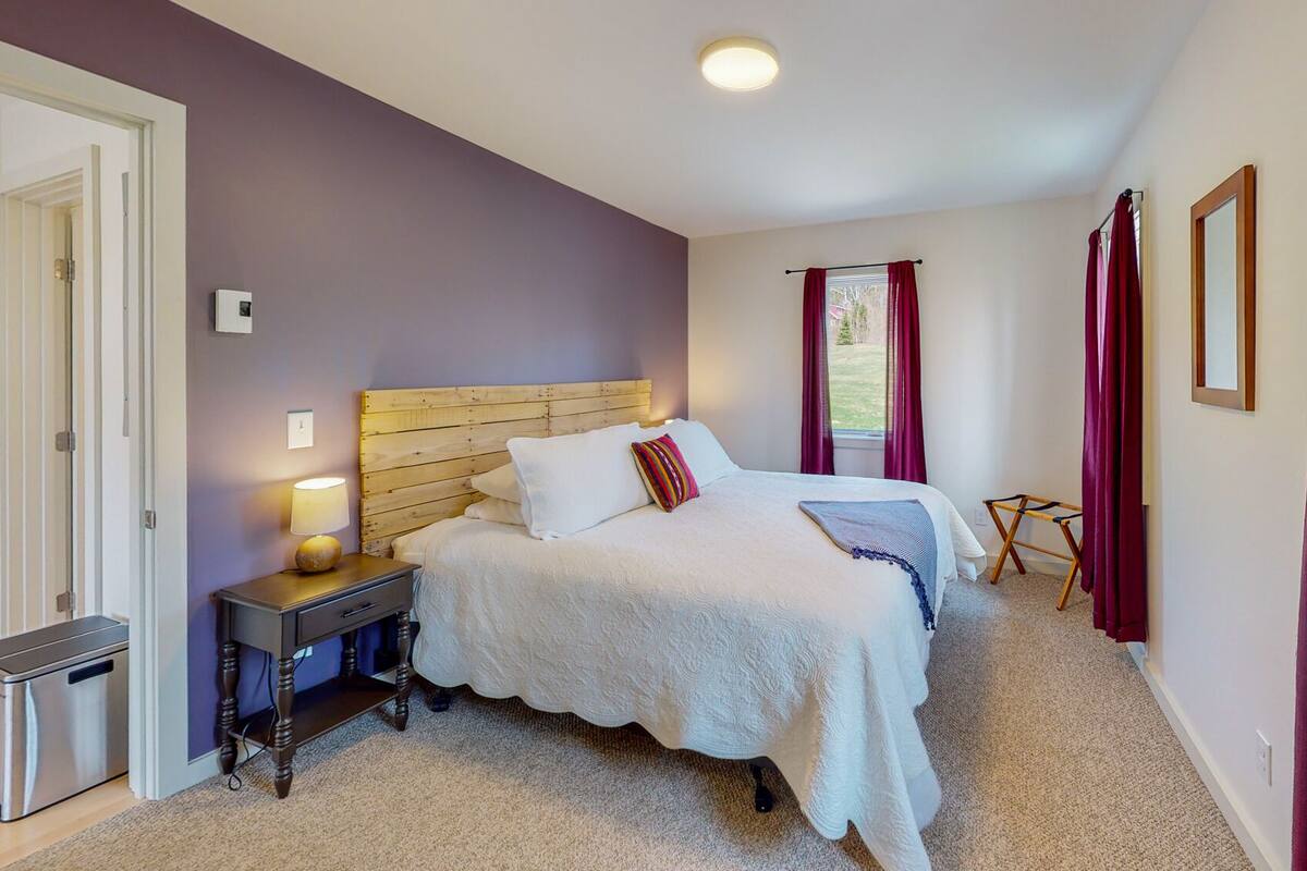 A bedroom with a wooden headboard, a bed with white linens, a bedside table with a lamp, and a window with red curtains.