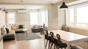 Een moderne woonkamer met een bank, een eettafel met stoelen, een kleine ronde tafel met een lamp en grote ramen die zorgen voor natuurlijk licht.