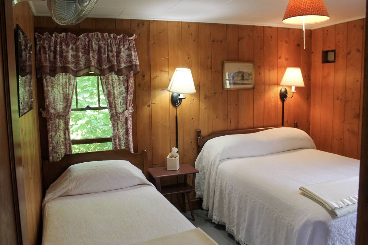 A room with two beds, wooden walls, a window with curtains, a bedside table with a lamp, and a fan.