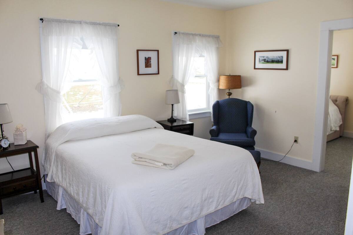 A hotel room with a bed, two bedside tables, a chair, and two windows with sheer curtains.