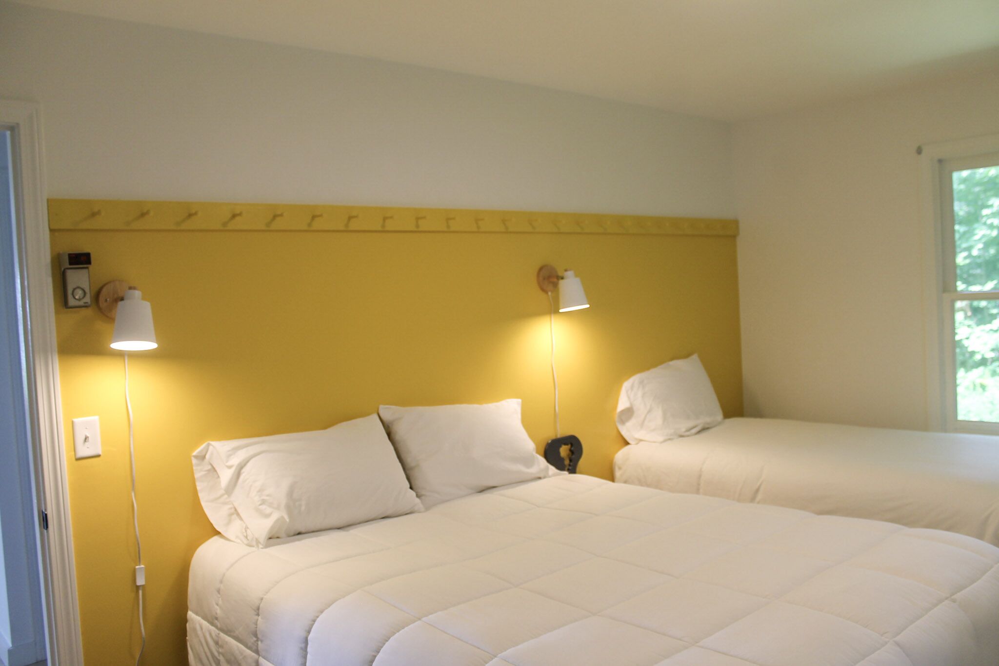 A room with two beds, each with white bedding and a headboard. There are two wall-mounted lamps on either side of the beds.