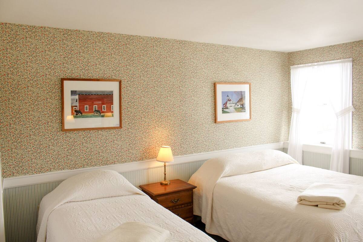 A hotel room with two beds, a nightstand, a lamp, and two framed pictures on the wall.
