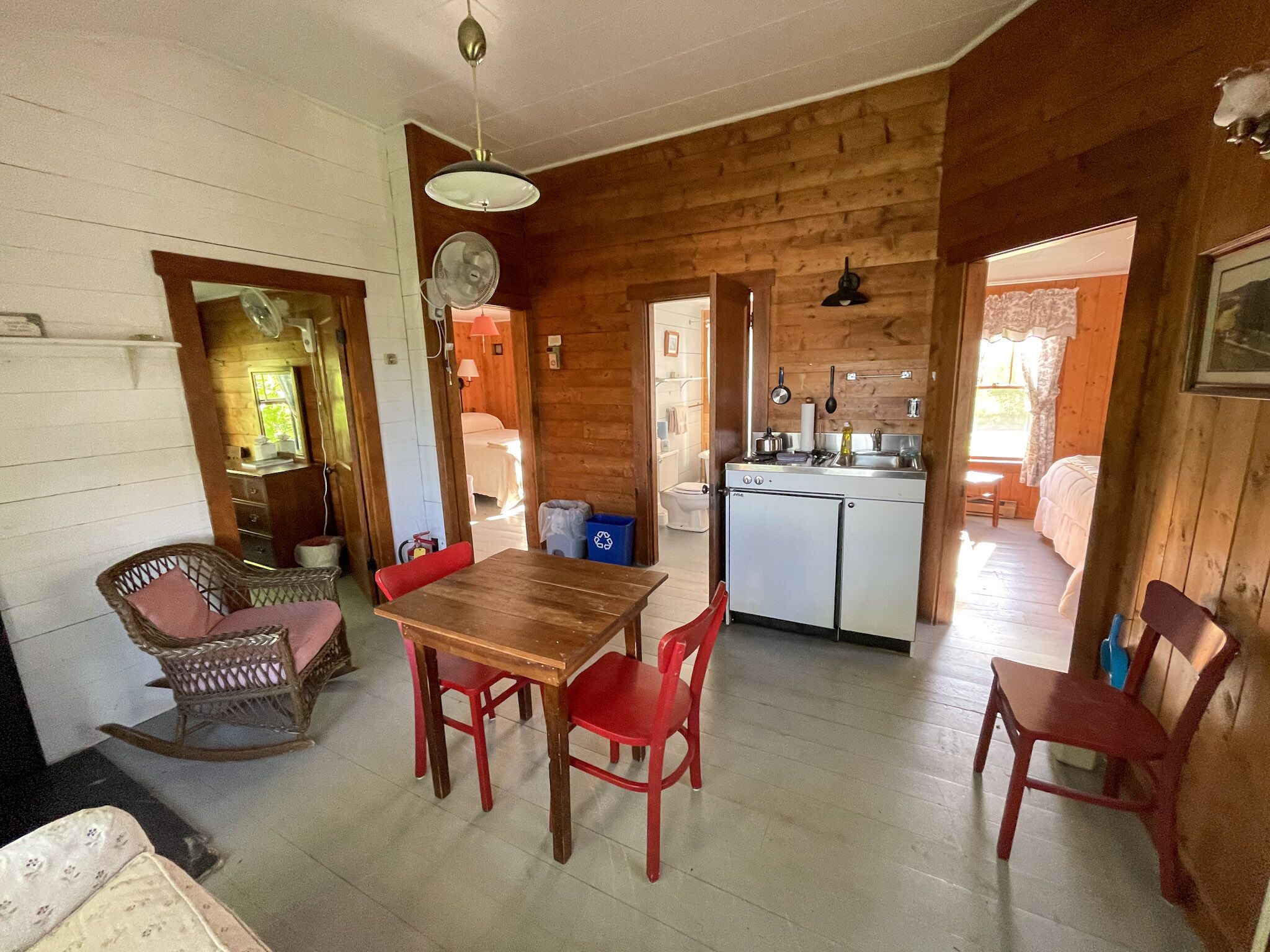 Una cabana acollidora amb interior de fusta, zona de dinar amb taula i cadires, petita cuina i un bany visible a través d'una porta oberta.